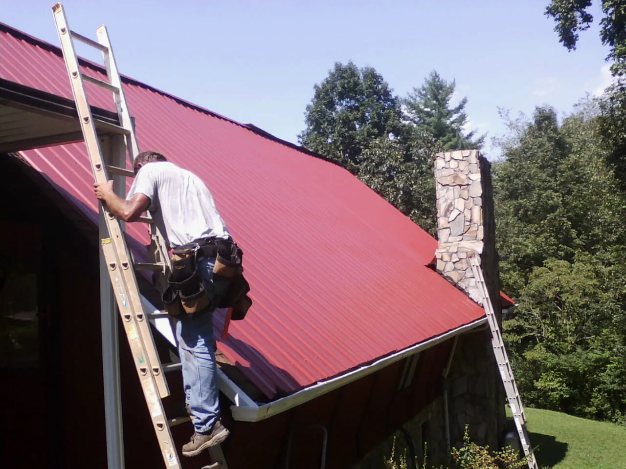 Roofer on a ladder beside a red metal roof and stone chimney — Soffit & Fascia Repair by Roofing Brothers in Noblesville